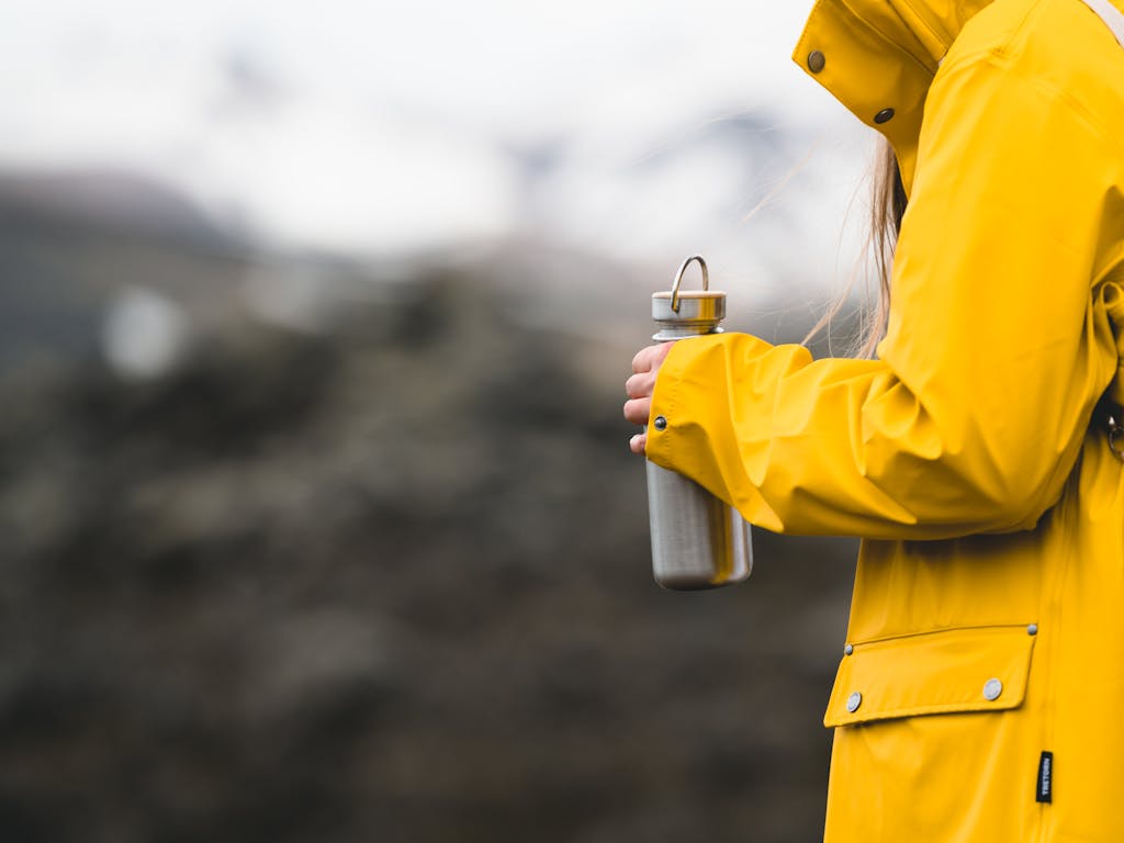 person in yellow rain jacket holding reusable water bottle on outdoor adventure – Sugar & Snip hydration blog