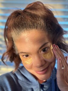 Woman applying under-eye patches during a morning skincare routine, touching her face in natural sunlight for a glowing skin look.