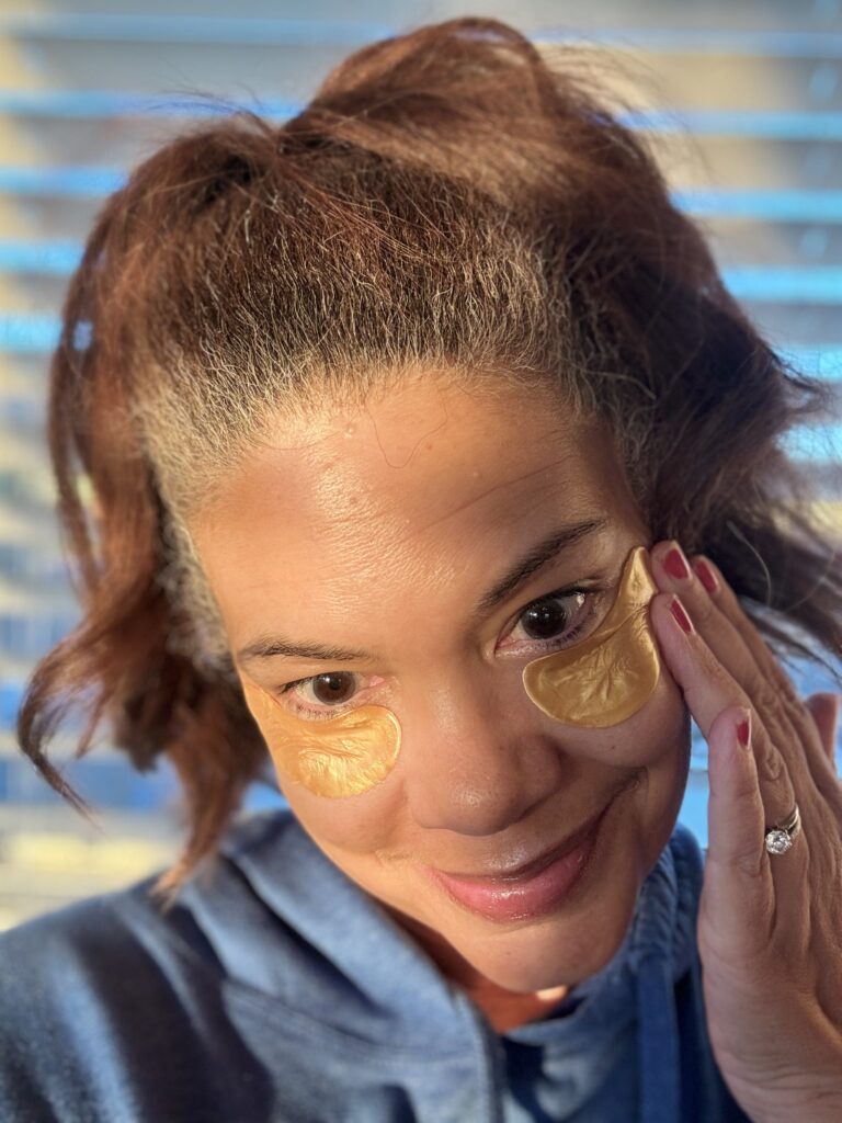 Woman applying under-eye patches during a morning skincare routine, touching her face in natural sunlight for a glowing skin look.