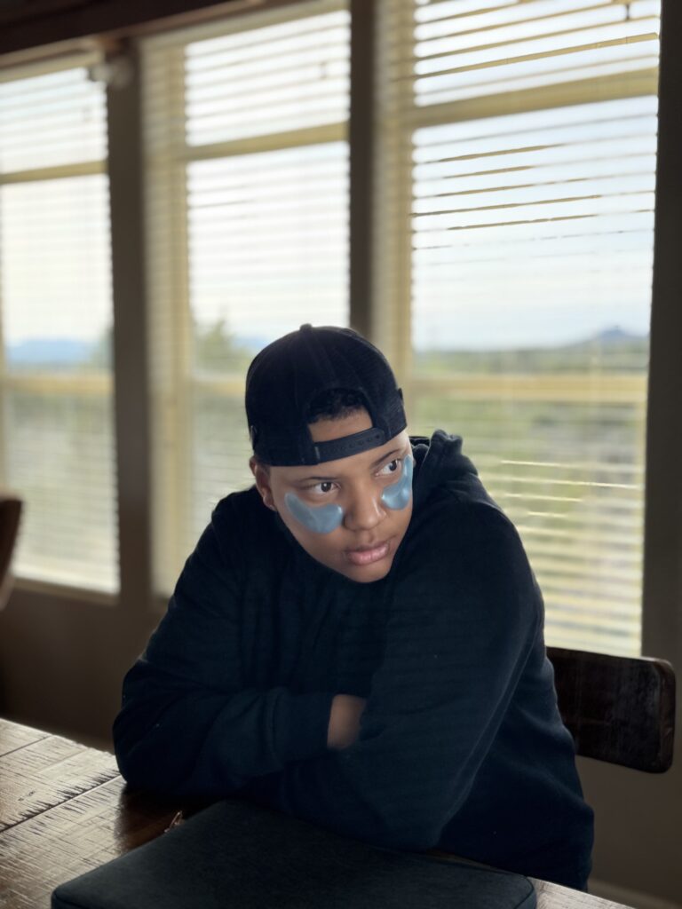 young black woman looking down with arms crossed wearing backward hat and blue eye patches for skin care morning routine
