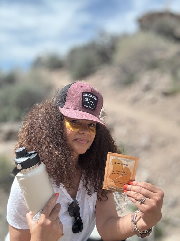 black woman wearing eye patches holding a bottle of water and  eye patches on a hike in a desert landscape