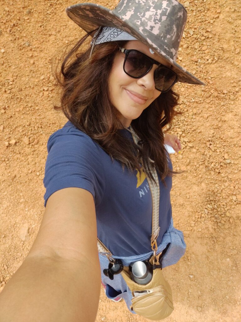 black woman wearing sunglasses and wide brim camo hat, shortsleeve tshirt on a summer hike