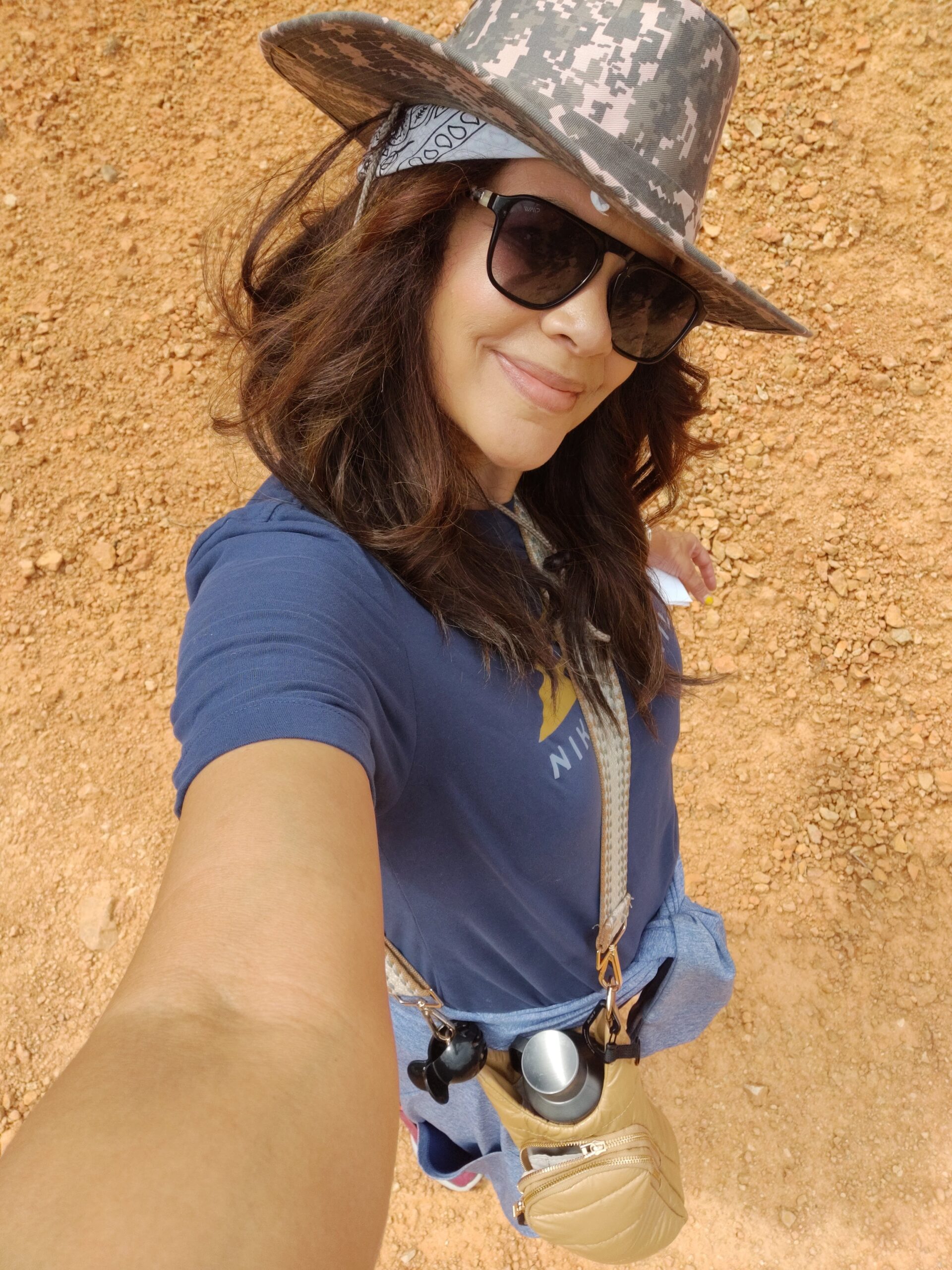 black woman wearing sunglasses and wide brim camo hat, shortsleeve tshirt on a summer hike