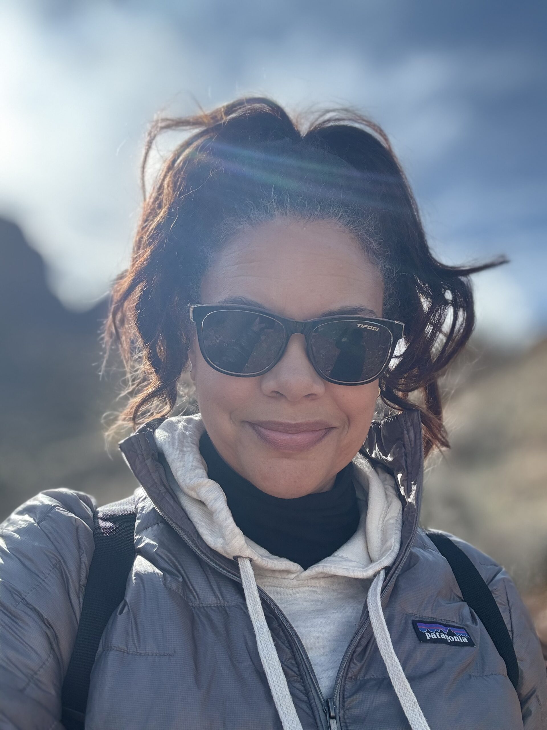 black woman wearing a patagonia jacket and sunglasses on a hike in Zion National Park with mountains and sun in the backgrouns