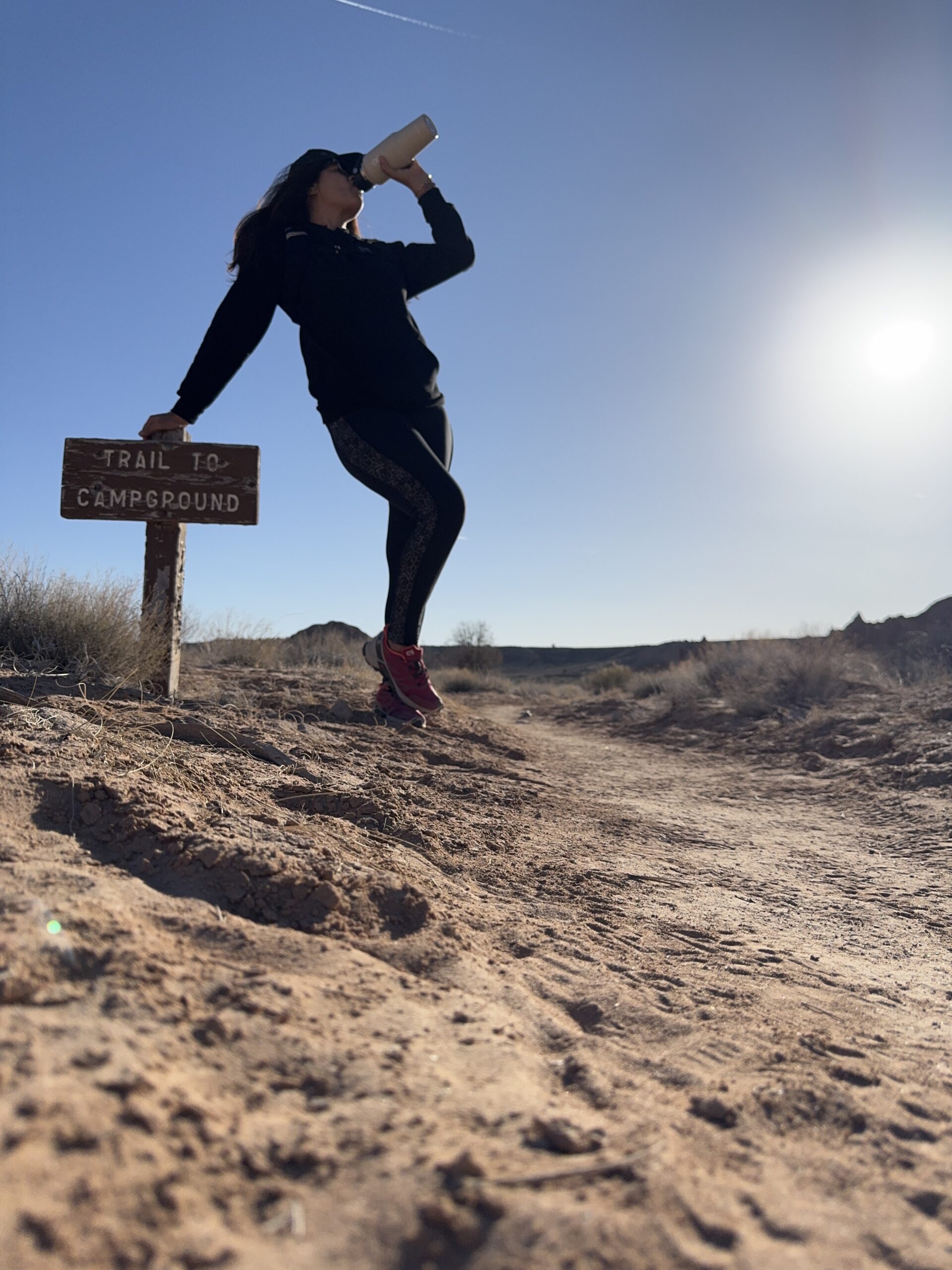 balck women hiking drinking water from a water bottle while leaning on a sign on a hike in a desert landscape