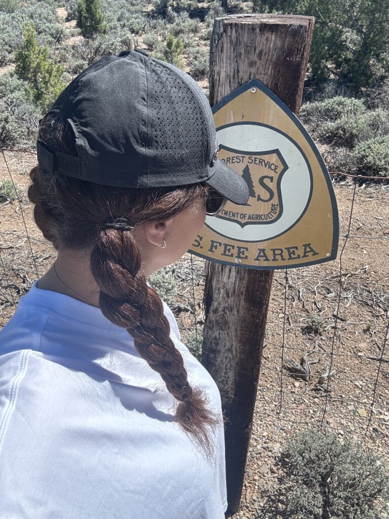 black woman with hair in braids wearing hat and looking and forest camping sign