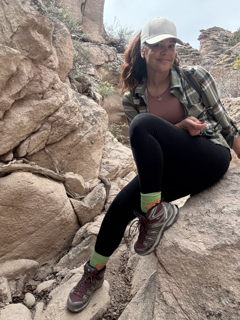 black woman sitting on rock in outdoorsy casual outfit with hat, plaid shirt and black leggings on a day hike.