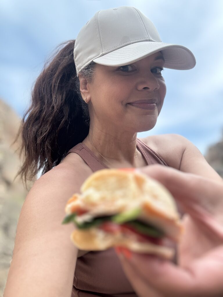 black woman in hat on a hike holding mini slider sandwich towards camera